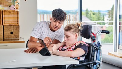 Ein junger Mann erklärt einer im Rollstuhl sitzenden Frau mit Beeinträchtigung etwas auf einem Tablet.