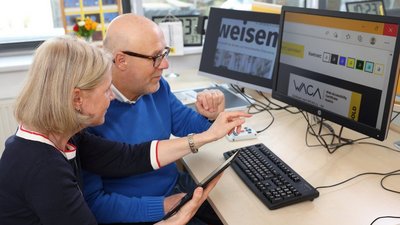 Eine Frau und ein Mann sitzen gemeinsam vor zwei Computerbildschirmen.