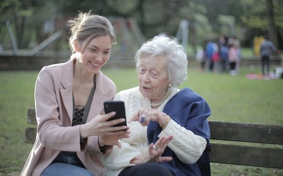 Eine junge weiße Frau zeigt einer älteren weißen Dame ihren Smartphone. Die junge Dame lächelt und die ältere Dame wirkt positiv überrascht.