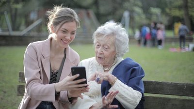 Eine junge weiße Frau zeigt einer älteren weißen Dame ihren Smartphone. Die junge Dame lächelt und die ältere Dame wirkt positiv überrascht.