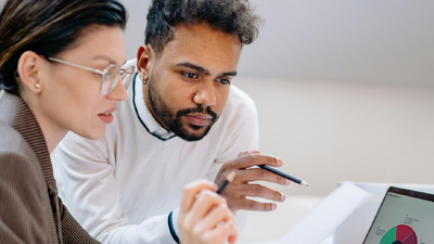 Eine Frau und ein Mann sitzen vor einem Laptop und besprechen etwas. Der Mann hält einen Bleistift in der Hand, die Frau einen Bleistift und ein Blatt Papier.