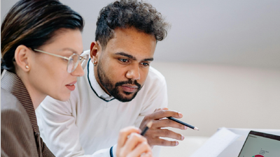 Eine Frau und ein Mann sitzen vor einem Laptop und besprechen etwas. Der Mann hält einen Bleistift in der Hand, die Frau einen Bleistift und ein Blatt Papier.