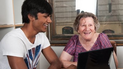 Ein junger Mann und eine ältere Dame vor einem Laptop sitzend.