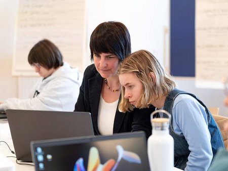 Frau erklärt einer anderen Frau etwas auf einem Laptop.