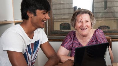 Ein junger Mann und eine ältere Dame vor einem Laptop sitzend.