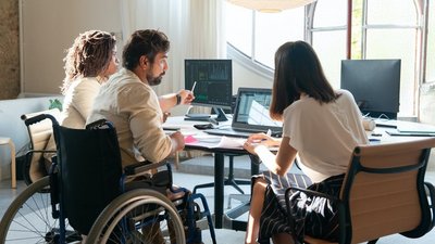 Drei Personen, die mittlere im Rollstuhl, sitzen mit dem Rücken zur Kamera um einen Schreibtisch.