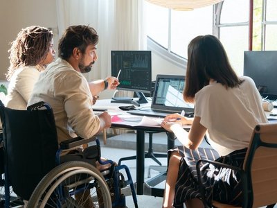 Drei Personen, die mittlere im Rollstuhl, sitzen mit dem Rücken zur Kamera um einen Schreibtisch.