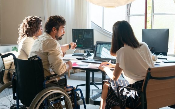 Drei Personen, die mittlere im Rollstuhl, sitzen mit dem Rücken zur Kamera um einen Schreibtisch.