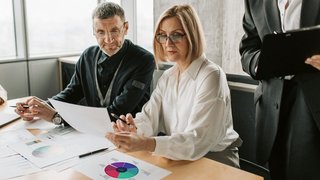 Drei Personen bei einem Tisch sitzend mit verschiedenen Dokumenten vor sich.
