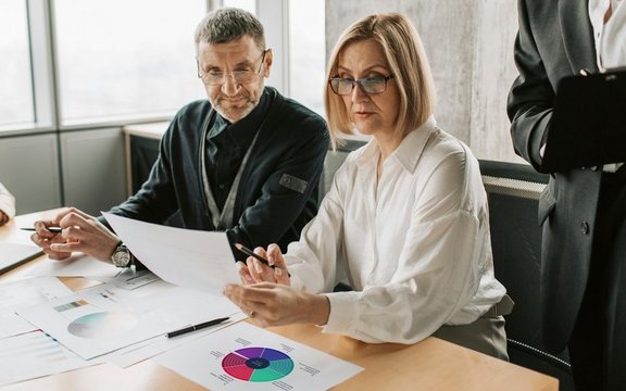 Drei Personen bei einem Tisch sitzend mit verschiedenen Dokumenten vor sich.