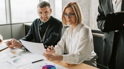 Drei Personen bei einem Tisch sitzend mit verschiedenen Dokumenten vor sich.
