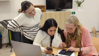 Zwei jüngere Frauen erklären einer älteren Dame etwas auf einem Smartphone, vor ihnen ein Laptop auf einem Tisch.